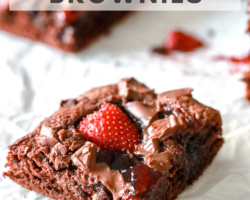 This is a side view of chewy and fudgey brownies. The brownie sits on a white piece of parchment paper. The brownie is topped with chopped chocolate, strawberry jam and sliced strawberries. More brownie squares are blurred in the background. Text overlay reads "amazingly fudgey strawberry brownies."