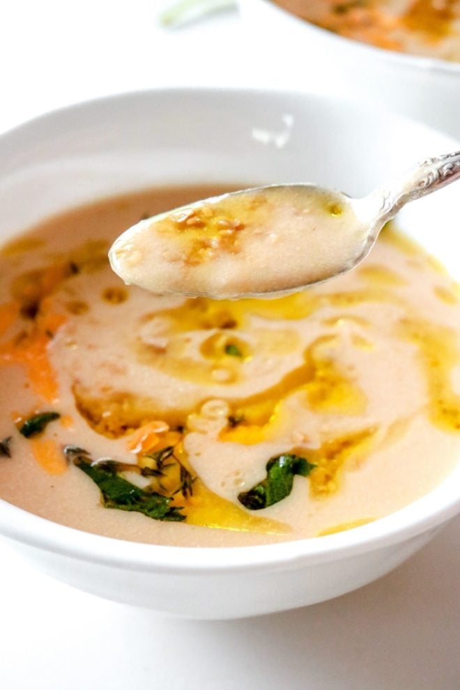 This is a closeup image of a soup pulling up a spoonful of soup from a bowl. The soup is topped with some garlic, oil, and sage.