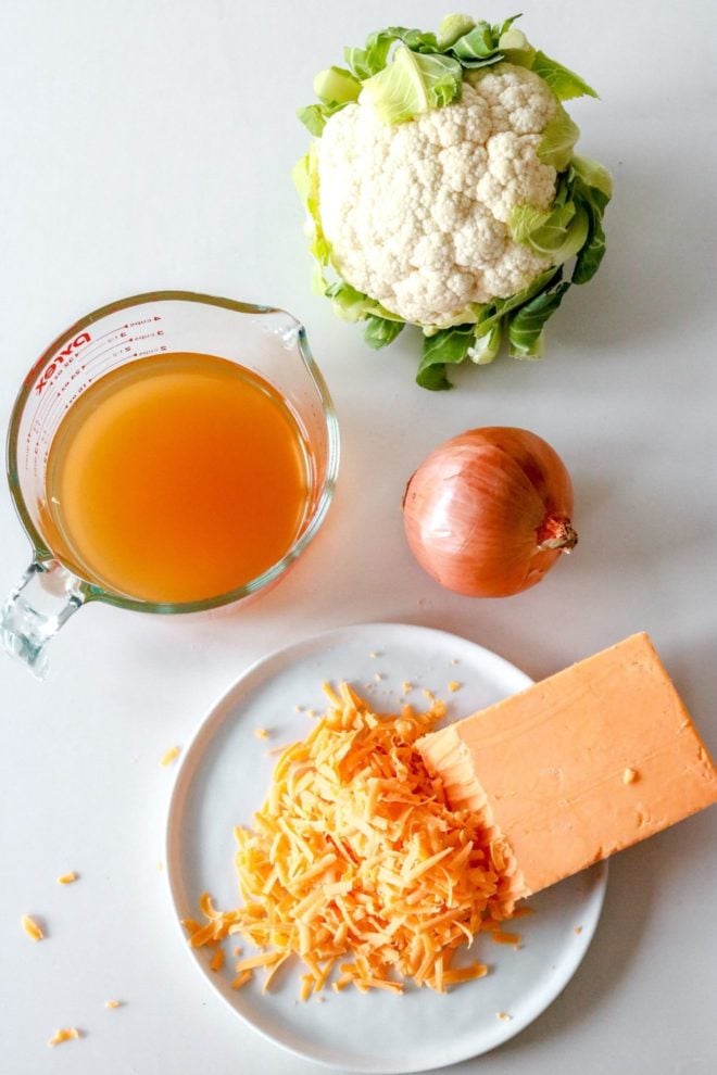 This is an overhead image of a cauliflower head, broth, and onion, and shredded cheddar cheese out on a white counter.