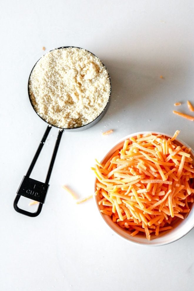 This is an overhead image of blanched almond flour in a measuring cup and shredded cheese in a bowl. The ingredients sit on a white counter.