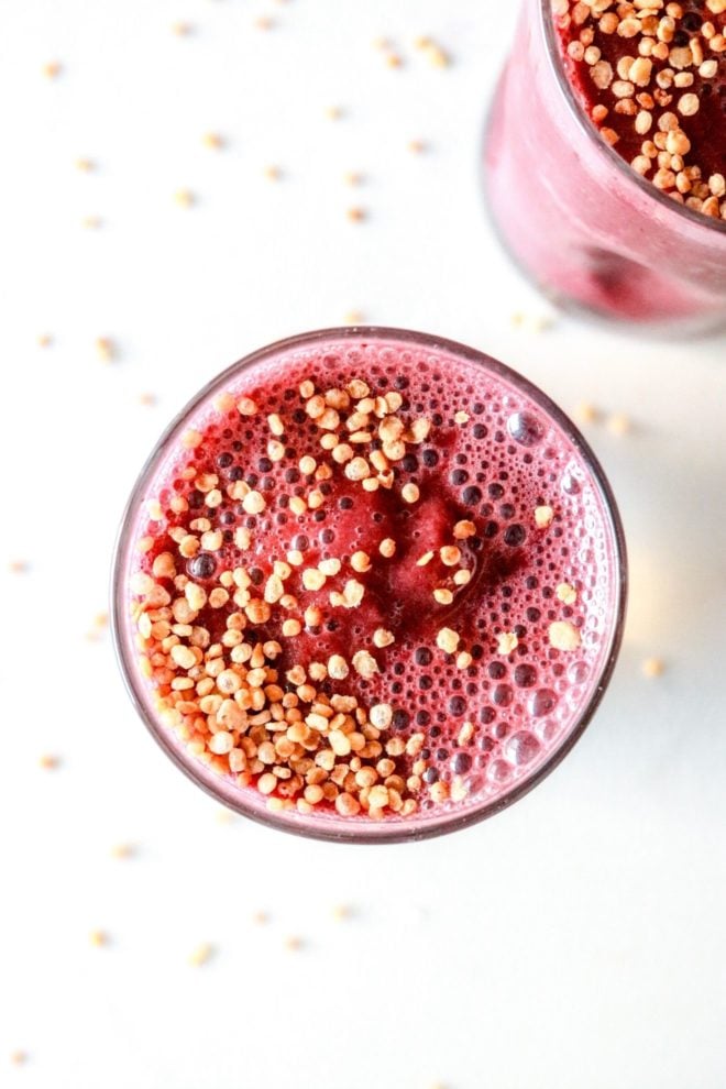 This is an overhead image of a glass on a white counter. Inside the glass is a cherry smoothie topped with puffed quinoa. More puffed quinoa is scattered on the white counter. Another glass filled with cherry smoothie is in the top right corner of the image.
