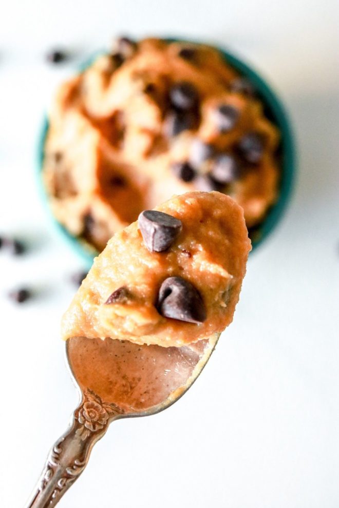 This is a close up of a spoon with chocolate chip cookie dough. A teal bowl with more cookie dough is on a white counter, blurred in the background.