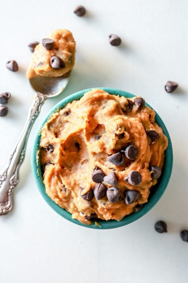 This is an overhead image of a small teal bowl filled with chocolate chip cookie dough. A spoon is scooping a bite of cookie dough and chocolate chips are scattered on the white counter around the bowl. A spoon with cookie dough is on the counter next to the bowl.