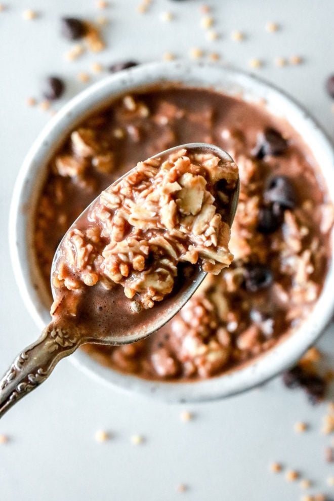 This is a closeup image of a spoon lifting up a bite of chocolate oats. Blurred in the background is a bowl sittng on a white counter. The bowl is filled with chocolate oats, chocolate chips, and puffed quinoa. More chocolate chips and puffed quinoa is scattered around the bowl on the counter.