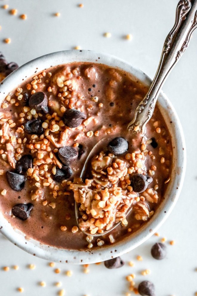 This is an overhead image of a bowl filled with chocolate liquid, chocolate chips, and puffed quinoa. A spoon is scooping up a bite of the oats. The bowl sits on a white counter.