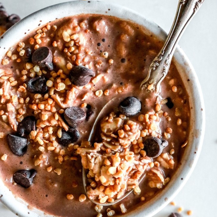 This is an overhead image of a bowl filled with chocolate oats, chocolate chips, and puffed quinoa. A spoon is scooping up a bite of the oats. The bowl sits on a white counter.