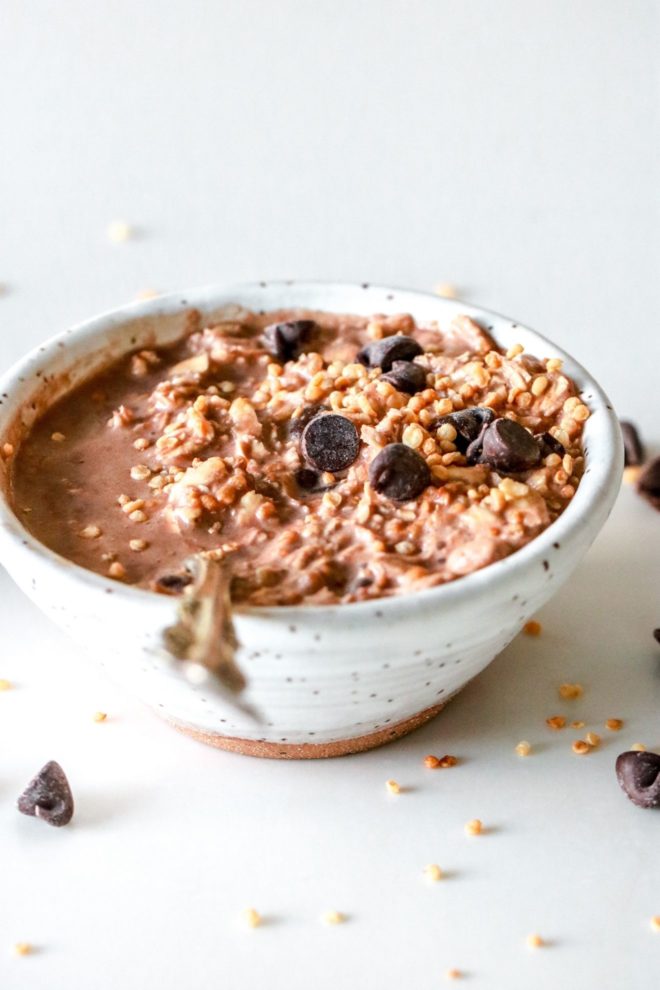 This is an image looking at a small white speckled bowl from the side. The bowl sits on a white counter and the bowl is filled with chocolate, chocolate chips, and puffed quinoa. A spoon is dipping into the bowl and leaning against the side of the bowl.