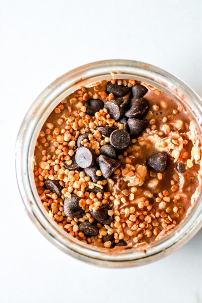 This is an overhead angle of a glass jar with overnight oats, chocolate chips, and puffed quinoa. The jar sits on a white counter.