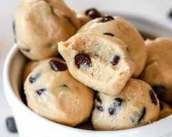 This is a close up side view of a small white bowl filled with raw cookie dough balls. The balls have chocolate chips in them and a few chocolate chips are scattered on the white counter around the bowl.