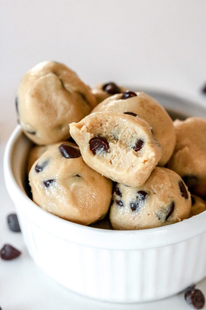 This is a close up side view of a small white bowl filled with raw cookie dough balls. The balls have chocolate chips in them and a few chocolate chips are scattered on the white counter around the bowl.