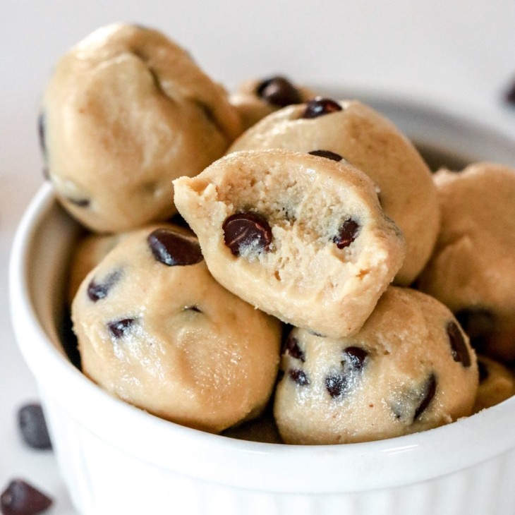 This is a close up side view of a small white bowl filled with raw cookie dough balls. The balls have chocolate chips in them and a few chocolate chips are scattered on the white counter around the bowl.