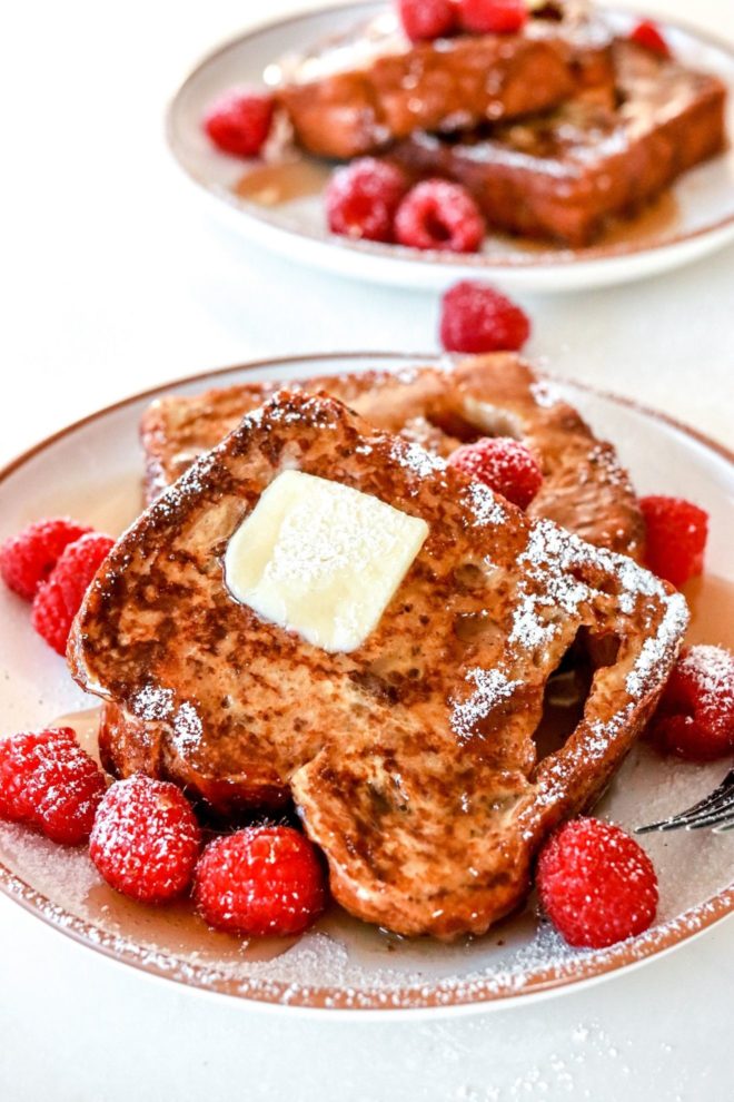 This is a side view of a plate of french toast. The french toast has butter, powdered sugar, syrup, and raspberries on top. The plate sites on a white counter and another plate with French toast is blurred in the background.