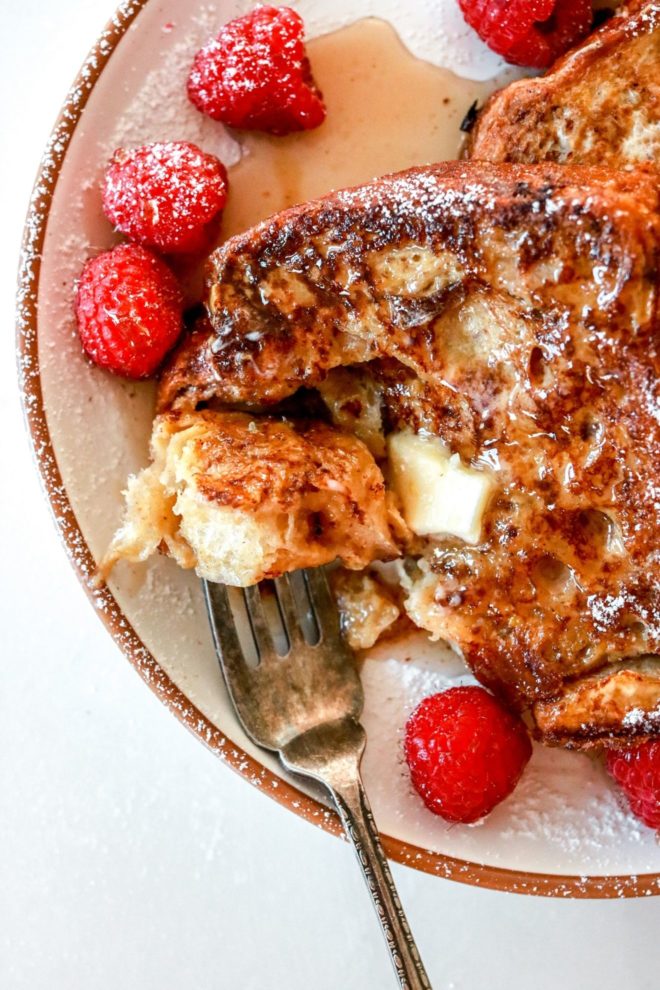This is an overhead image of french toast on a plate with raspberries, powdered sugar, syrup, and butter. The plate sits on a white counter. A fork is piercing a bite of french toast and is leaning against the side of the plate.