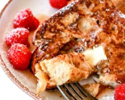 This is a side image of a plate with french toast and raspberries on it. A fork is piercing a bite of french toast and is laying on the side of the plate.