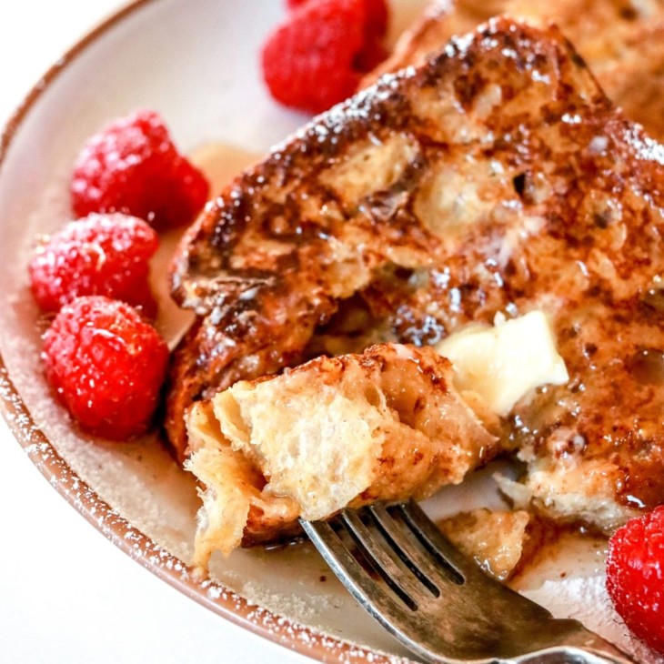 This is a side image of a plate with french toast and raspberries on it. A fork is piercing a bite of french toast and is laying on the side of the plate.