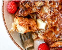 This is an overhead image of french toast on a plate with raspberries, powdered sugar, syrup, and butter. The plate sits on a white counter. A fork is piercing a bite of french toast and is leaning against the side of the plate. Text overlay reads "healthy french toast dairy free & easy to make!"