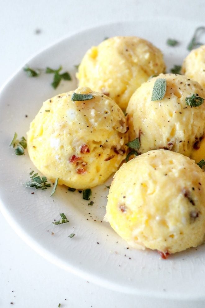 This is a side image of egg bites on a white plate. The egg bites have pieces of bacon in them and are topped with chopped sage and ground pepper.