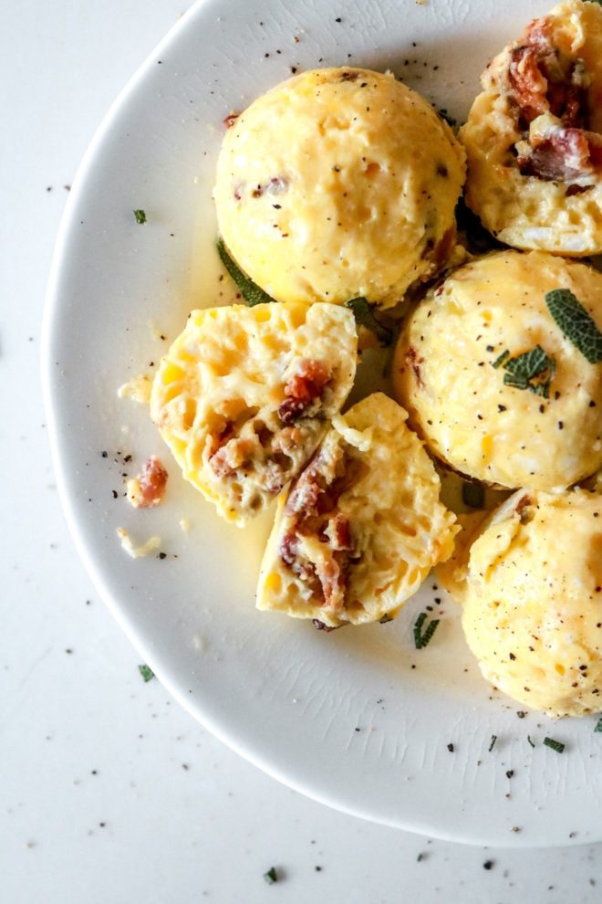 This is an image of a white plate with egg bites on it. One egg bite is cut open and cheese and bacon is inside. The egg bites are topped with chopped sage and ground pepper.