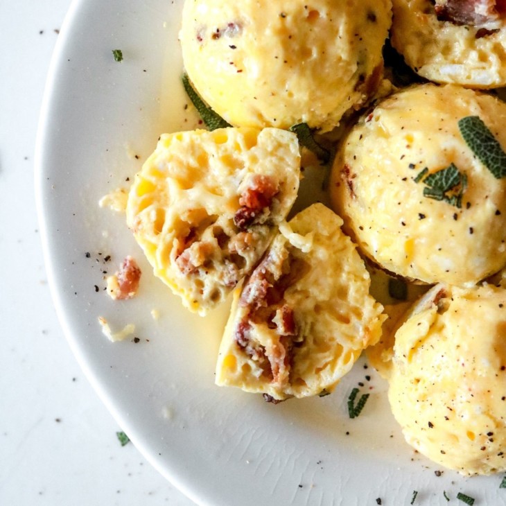 This is an image of a white plate with egg bites on it. One egg bite is cut open and cheese and bacon is inside. The egg bites are topped with chopped sage and ground pepper.
