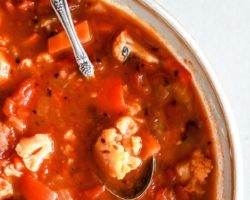 This is an overhead image of a bowl of vegetable soup. The soup has a red broth with chunky vegetables and a spoon scooping a cauliflower floret and other chopped vegetables.