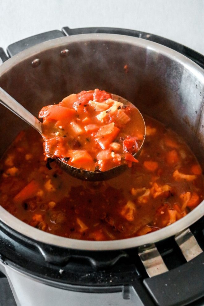 This is an image looking into an instant pot with vegetable soup inside. A ladle is lifting up a steaming scoop of vegetable soup.