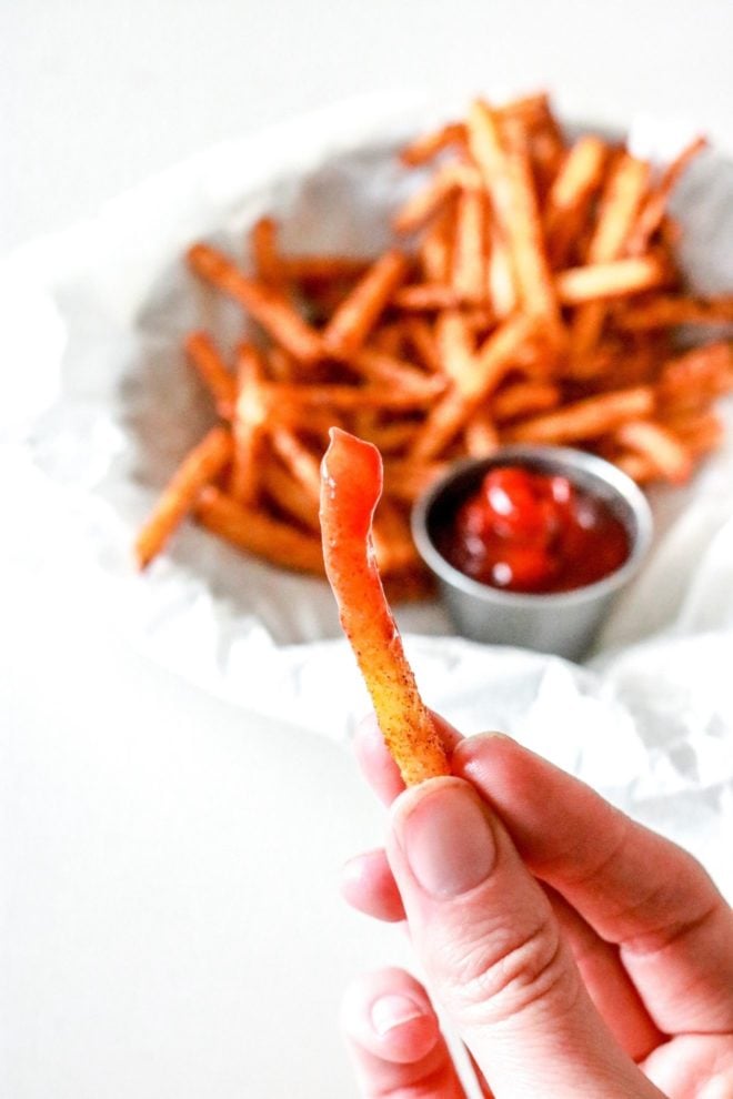 This is an image of a hand holding a jicama fry dipped in ketchup. A plate of jicama fries with a small bowl of ketchup is blurred in the background.