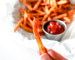 This is an image of a hand holding a jicama fry dipped in ketchup. A plate of jicama fries with a small bowl of ketchup is blurred in the background. Text overlay reads "crispy jicama fries".