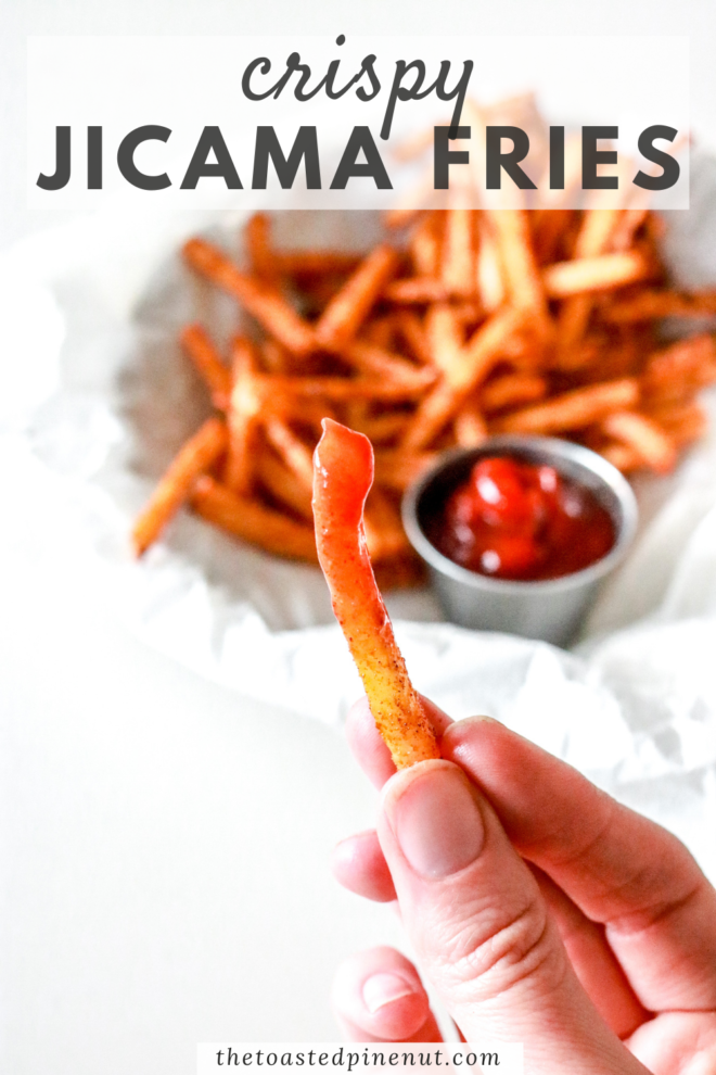 This is an image of a hand holding a jicama fry dipped in ketchup. A plate of jicama fries with a small bowl of ketchup is blurred in the background. Text overlay reads "crispy jicama fries".