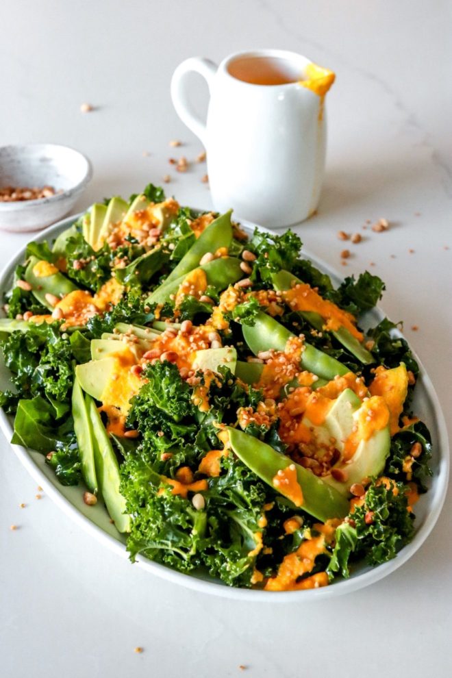 This is a side view of a platter of kale salad sitting on a white counter. The salad is topped with snow peas, avocado, and toasted nuts. A small bowl of nuts is off to the side. A small pitcher of ginger carrot dressing is off to the side next to the salad platter.