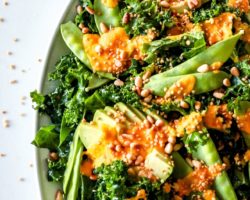 This is a close up overhead image of a white platter with kale salad with avocado, snow peas, an orange dressing, toasted pine nuts, and puffed quinoa.