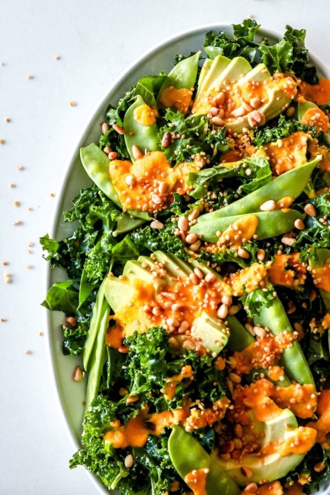 This is a close up overhead image of a white platter with kale salad with avocado, snow peas, an orange dressing, toasted pine nuts, and puffed quinoa.