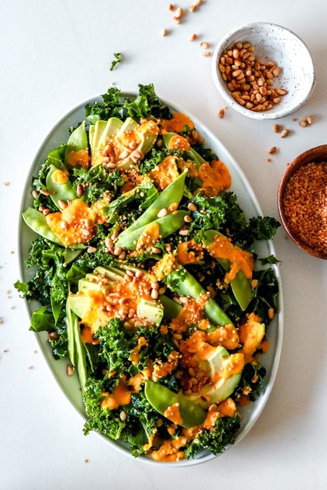 This is an overhead image of a white platter on a white counter. On the platter is a kale salad with snow peas, avocado, an orange dressing, and toasted nuts. Small bowls with nuts and quinoa are off to the side, next to the platter.