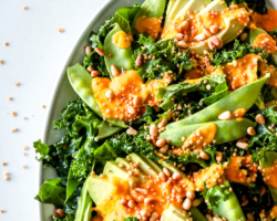 This is a close up overhead image of a white platter with kale salad with avocado, snow peas, an orange dressing, toasted pine nuts, and puffed quinoa. Text overlay reads "kale avocado salad with carrot ginger dressing."