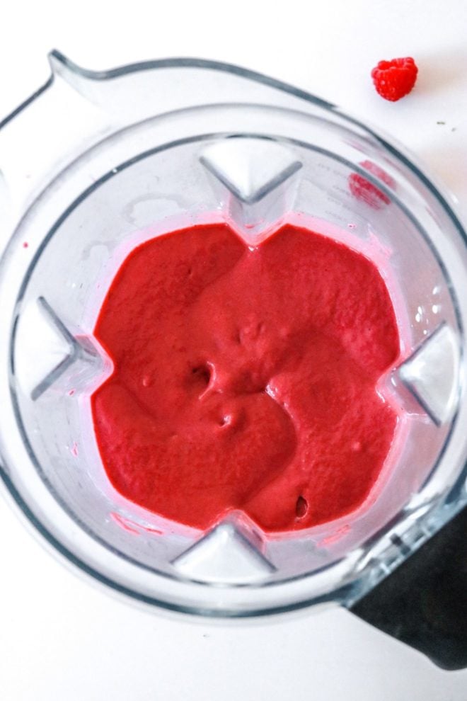 This is an overhead image of a blender filled with a raspberry smoothie. A couple fresh raspberry are on the white counter around the blender.