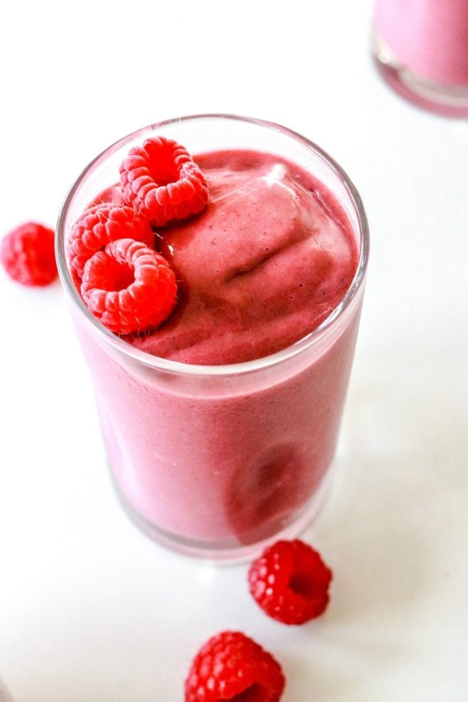 This is an overhead view of a raspberry smoothie in a glass on a white counter. Another glass filled with the smoothie is in the top right corner of the image and fresh raspberries are on the counter around the glass.