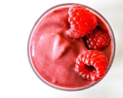 This is an overhead image of a raspberry smoothie in a glass on a white counter. Fresh raspberries are at the top and bottom of the image.