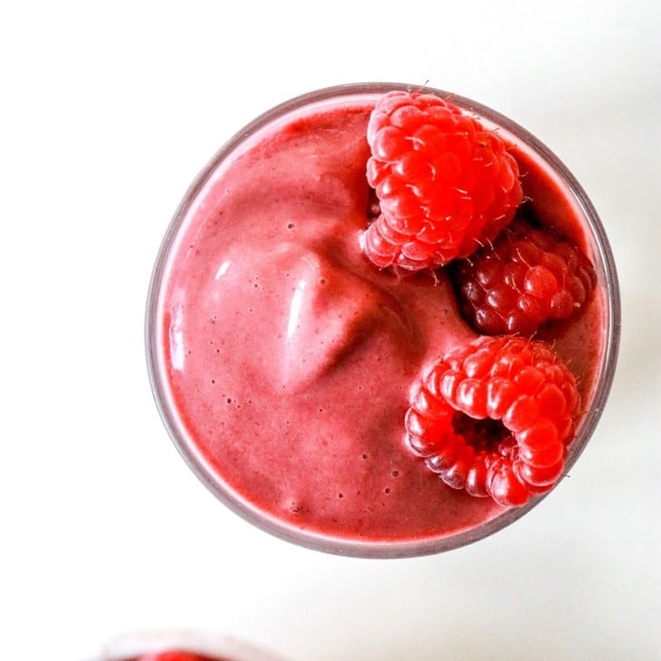 This is an overhead image of a raspberry smoothie in a glass on a white counter. Fresh raspberries are at the top and bottom of the image.