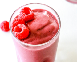 This is an overhead view of a raspberry smoothie in a glass on a white counter. Another glass filled with the smoothie is in the top right corner of the image and fresh raspberries are on the counter around the glass. Text overlay reads "6 ingredient raspberry smoothie."