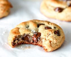 This is a side view of a chocolate chip cookie with a bite taken out of it. The Cookie sits on a white piece of parchment paper with more cookies blurred in the background.