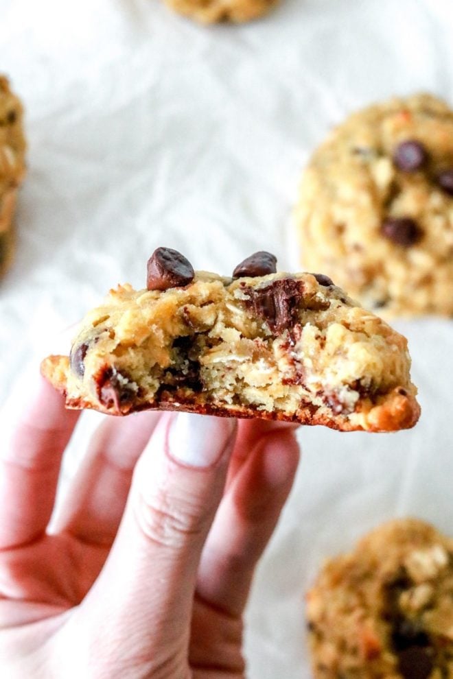 A hand is holding a chocolate chip oatmeal cookie with a bite taken out of it. More cookies are blurred in the background sitting on a white piece of parchment paper.