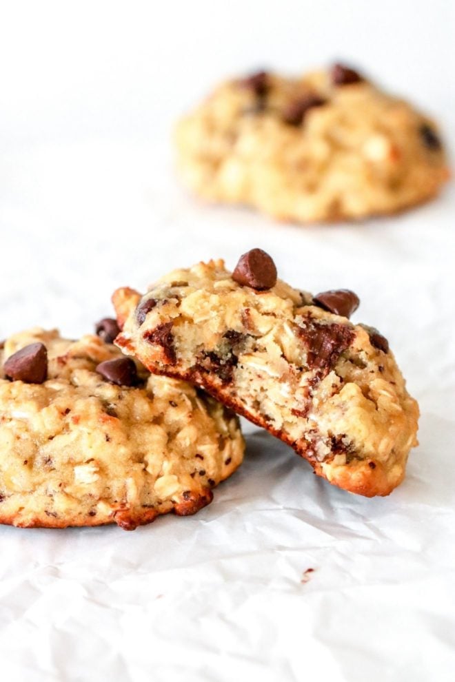 This is a side view of an oatmeal chocolate chip cookie with a bite taken out of it. The cookie leans against another cookie. The cookie sits on a white piece of parchment paper. Another cookie is blurred in the background.