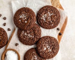 This is an overhead view of a cutting board with a piece of parchment paper on top of a bunch of chocolate cookies. The cookies have crinkle tops and salt sprinkled on top. A small wood bowl filled with flakey salt and chocolate chips are next to the cookies.
