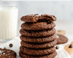 This is a stack of chocolate cookies on a cutting board with parchment paper. More cookies are around the stack of cookies and a glass of milk is blurred in the background. Text overlay reads "chewy gluten free brownie cookies."