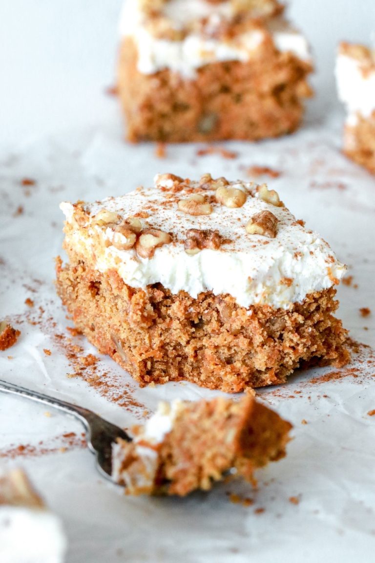 This is a side view of a carrot cake bar with cream cheese frosting. The carrot cake is topped with chopped walnuts and a sprinkle of cinnamon. A fork is taking a bite out of the cake and laying in the front. The cake bar is on a piece of white parchment paper with more bars blurred din the background.