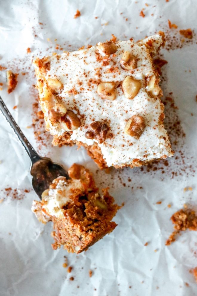 This is an overhead view of a carrot cake bar with cream cheese frosting. The carrot cake is topped with chopped walnuts and a sprinkle of cinnamon. A fork is taking a bite out of the cake and laying in the front. The cake bar is on a piece of white parchment paper.