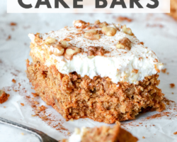 This is a side view of a carrot cake bar with cream cheese frosting. The carrot cake is topped with chopped walnuts and a sprinkle of cinnamon. A fork is taking a bite out of the cake and laying in the front. The cake bar is on a piece of white parchment paper with more bars blurred din the background. Text overlay reads "healthy oat flour carrot cake bars"