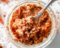 This is an overhead image of a glass jar with carrots, oats, pecans, and milk in it. A spoon full of oatmeal is leaning against the side of the jar. More shredded carrot and nuts are on the counter around the jar.