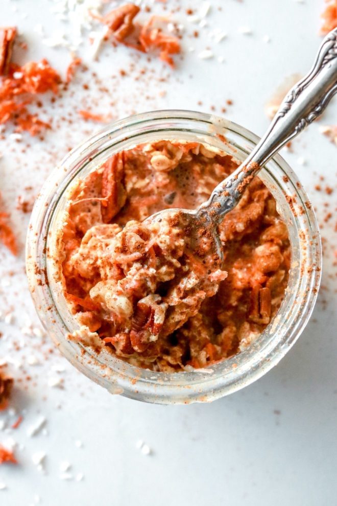 This is an overhead image of a glass jar with carrots, oats, pecans, and milk in it. A spoon full of oatmeal is leaning against the side of the jar. More shredded carrot and nuts are on the counter around the jar.