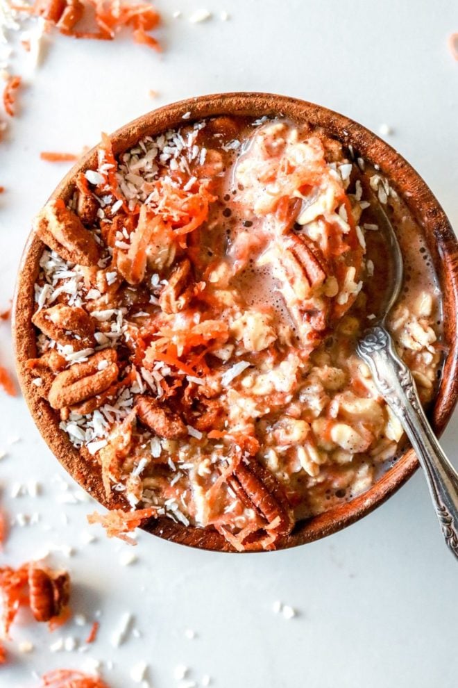 This is an overhead image of a small wood bowl with carrots, oats, pecans, and shredded coconut in it. A spoon full of oatmeal is leaning against the side of the bowl. More shredded carrot and nuts are on the counter around the bowl.