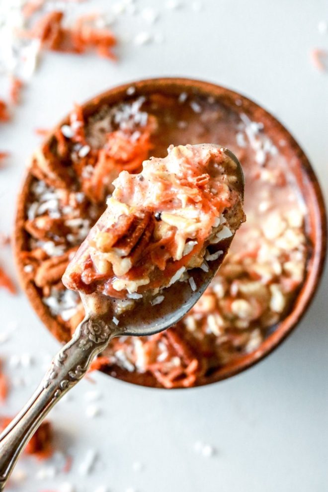 This is an overhead image of a small wood bowl with shredded carrots, oats, pecans, and shredded coconut in it. A spoon full of oatmeal is in focus above the bowl.